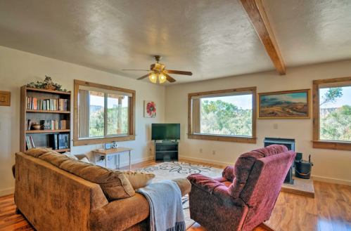 Cozy living room with a sofa, armchair, TV, and large windows showcasing greenery outside. Warm, inviting atmosphere.